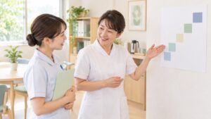明るいスタッフルームで、先輩の看護師が壁に貼った階段状の図を指しながら、若い看護師に笑顔で説明している。