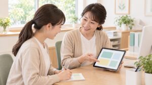 受付の机で、先輩スタッフが新しく入ったスタッフにタブレットの画面を見せながら手順を説明し、新人が笑顔でメモを取っている。